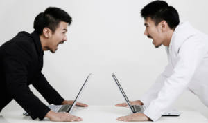 Two businessmen standing over their computers yelling at one another.